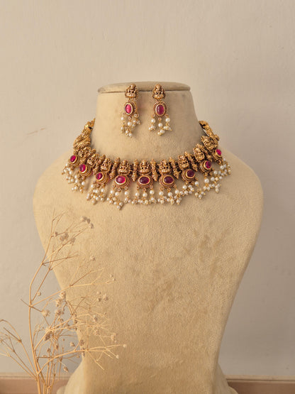 Sonali Antique Gold Temple Choker Set displayed on a beige mannequin, featuring traditional goddess motifs embedded with pink ruby stones and bordered with delicate cascading pearl drops, accompanied by matching jhumka-style earrings by Naitri.in