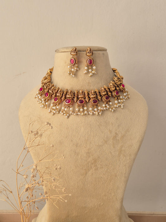 Sonali Antique Gold Temple Choker Set displayed on a beige mannequin, featuring traditional goddess motifs embedded with pink ruby stones and bordered with delicate cascading pearl drops, accompanied by matching jhumka-style earrings by Naitri.in