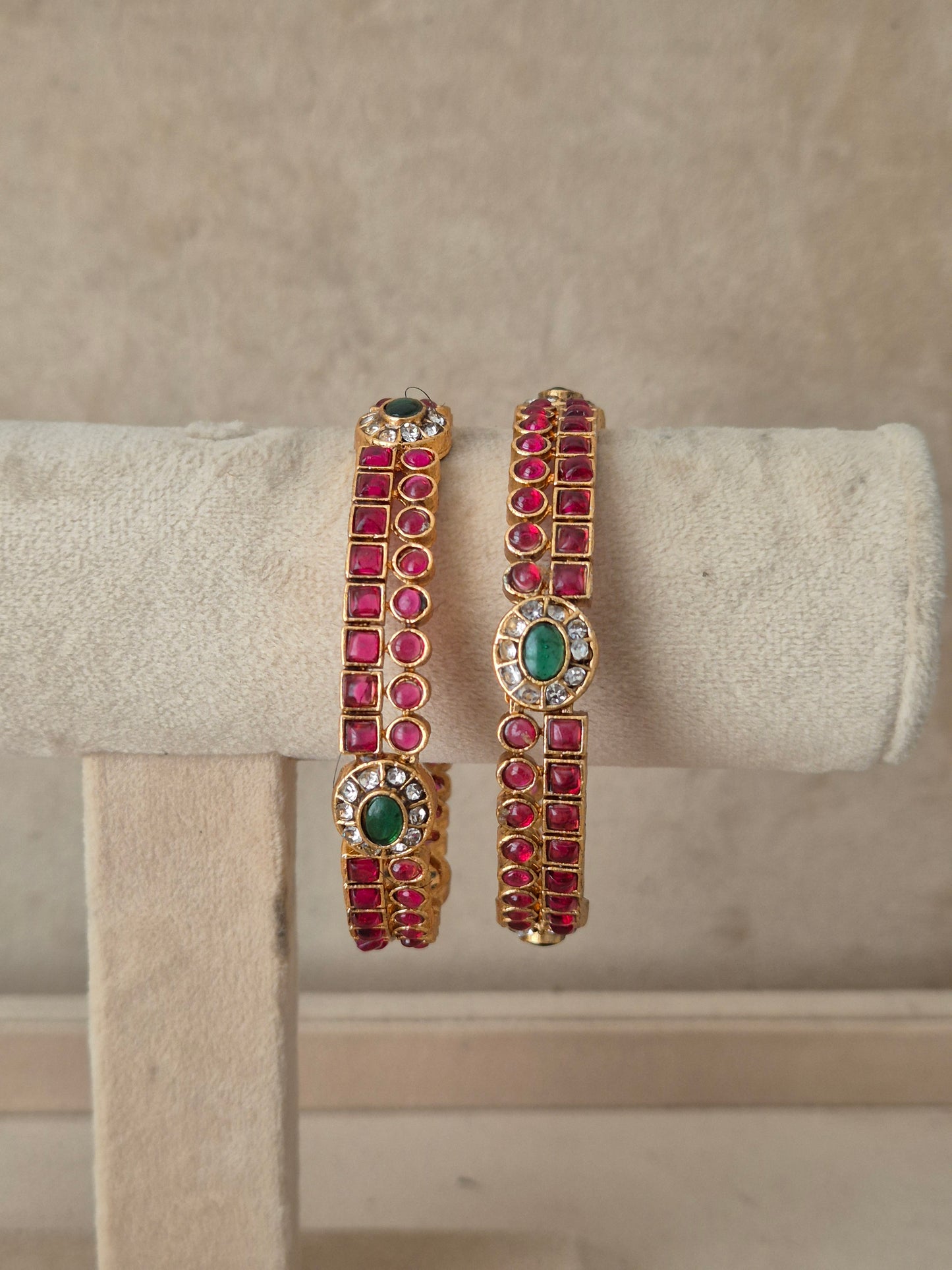 A pair of gold-plated cuff bracelets decorated with rows of pinkish-red round gemstones and green oval center stones, by naitri.in