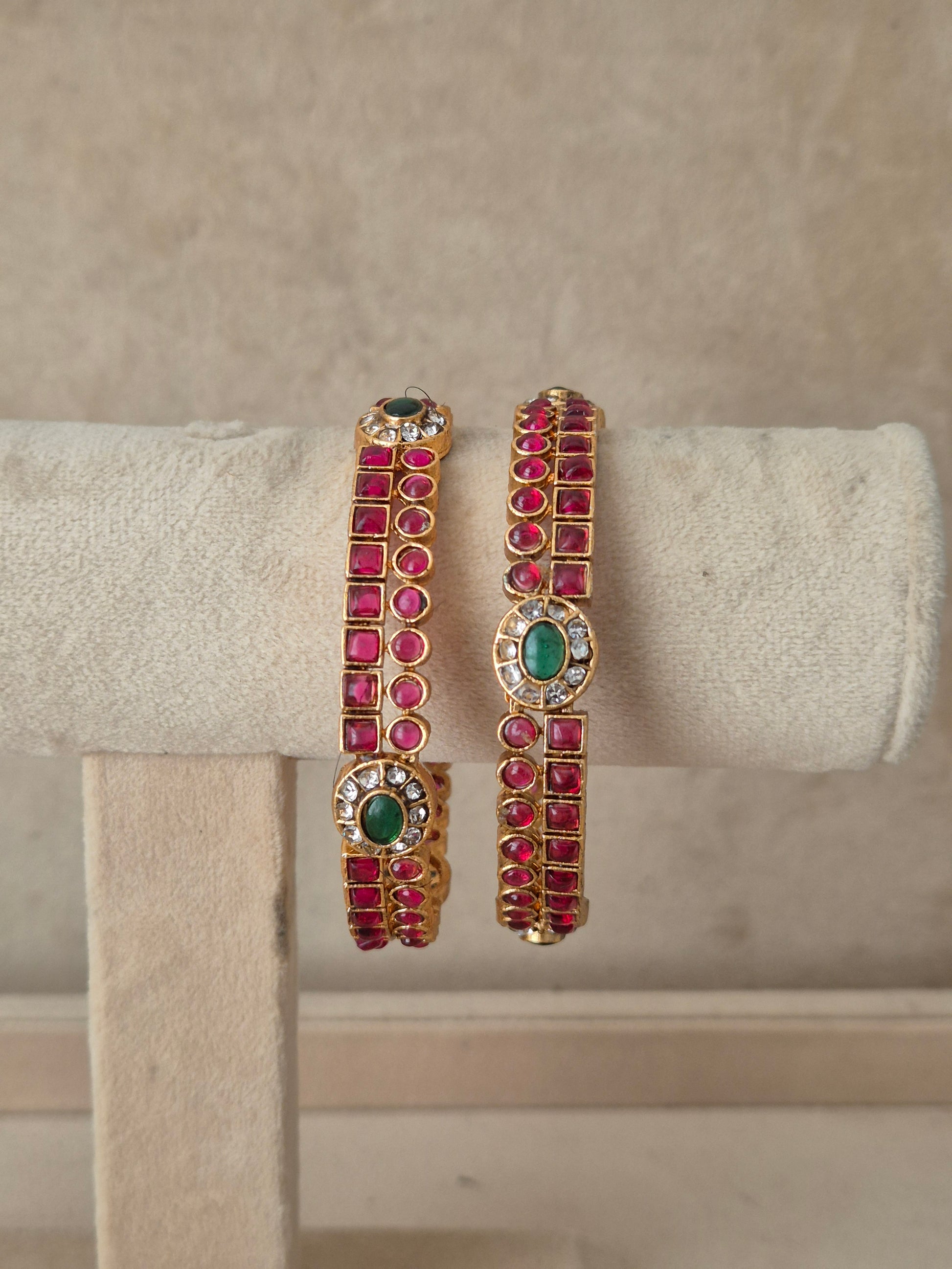 A pair of gold-plated cuff bracelets decorated with rows of pinkish-red round gemstones and green oval center stones, by naitri.in
