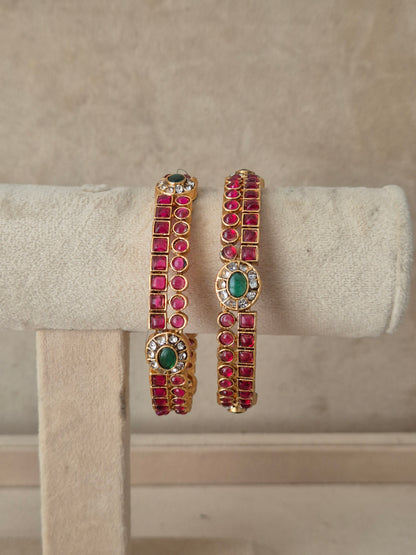 A pair of gold-plated cuff bracelets decorated with rows of pinkish-red round gemstones and green oval center stones, by naitri.in