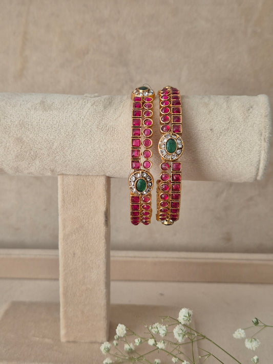 Pair of gold-plated bangles studded with square-cut pink stones and a central oval green gemstone by naitri.in