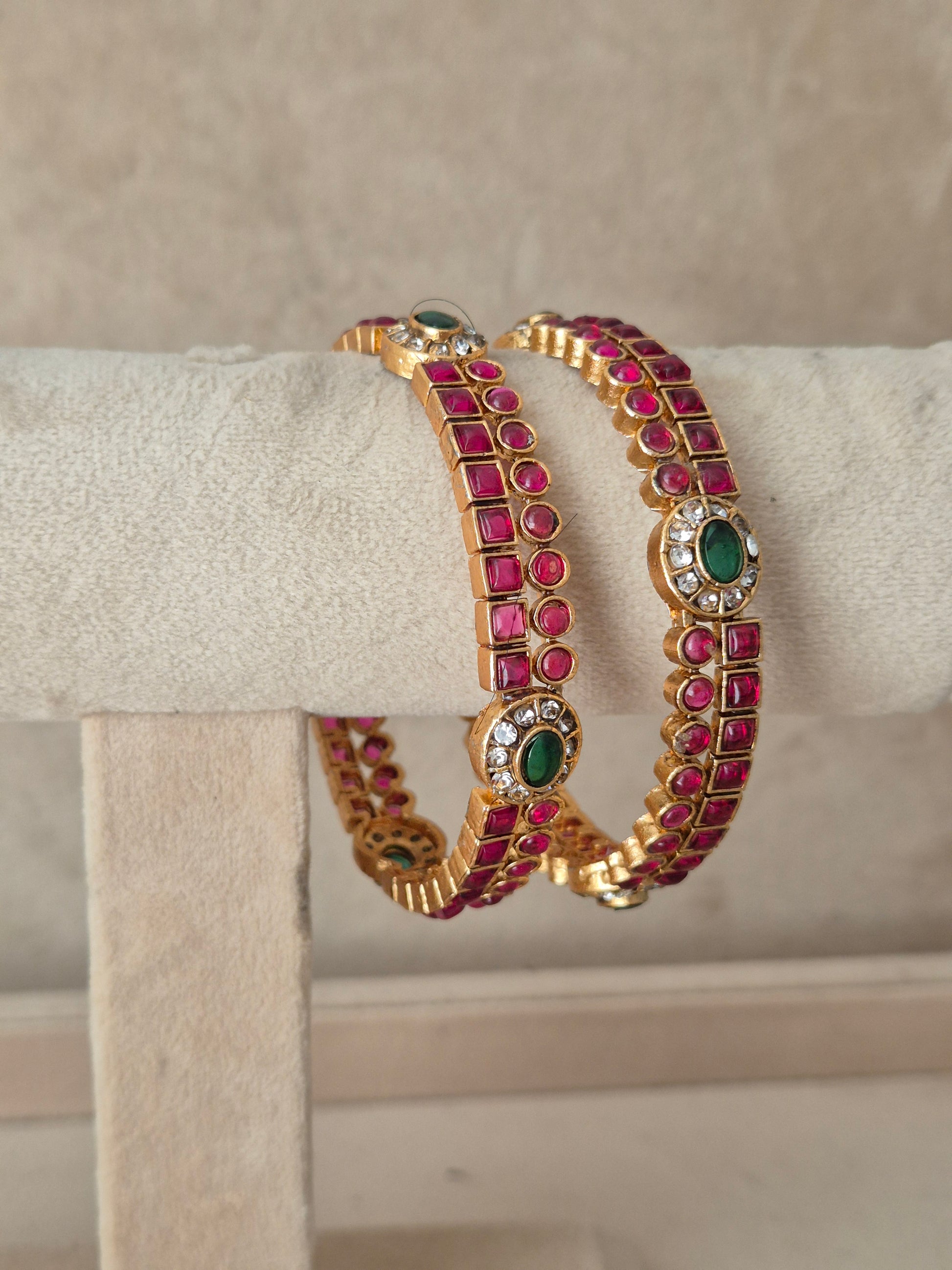 Two gold-plated bangles decorated with rows of pinkish-red round stones and green Traditional Indian bridal bangles adorned with pink gemstones and an oval emerald green accent stone oval stones, by naitri.in