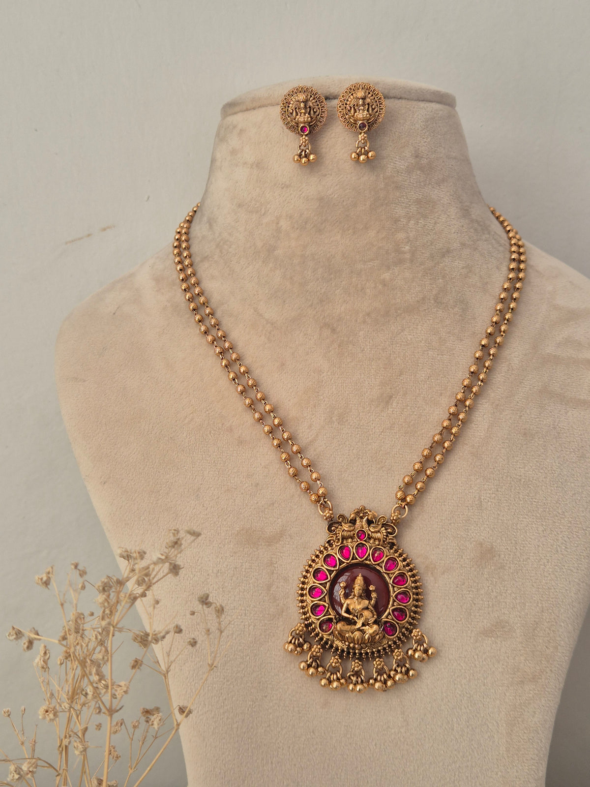 Traditional gold long chain temple necklace with a circular pendant featuring a goddess motif surrounded by pink stones and small gold drop tassels with matching earrings by naitri.in