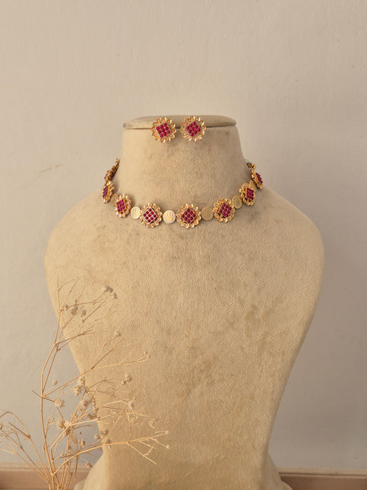 Titli Ruby Floral Choker Set displayed on a neutral beige mannequin, featuring a delicate arrangement of vibrant ruby-pink stone floral motifs along a golden chain, accompanied by matching stud earrings by Naitri.in