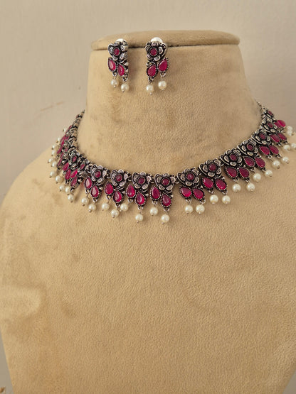 Pair of oxidized silver floral motif choker necklace with matching redish pink stone earrings by Naitri.in