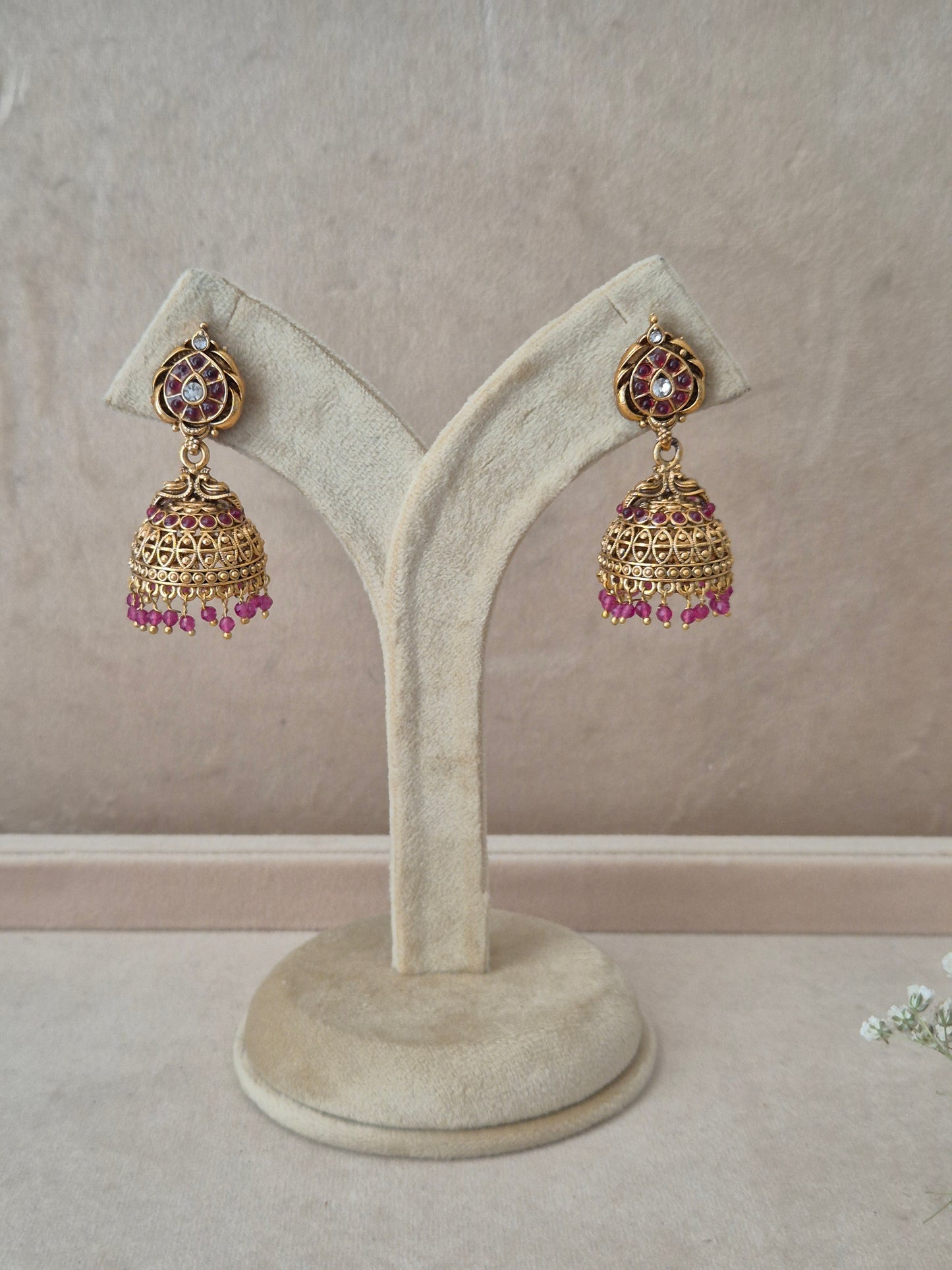 Pair of gold-plated Urjita Jhumka earrings hanging on a beige velvet display stand, featuring circular teardrop-accented studs and bell-shaped drops adorned with a fringe of dangling pink beads by naitri.in