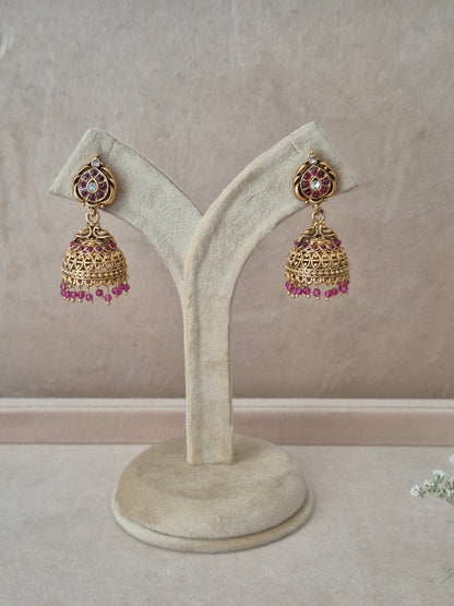 Pair of gold-plated Urjita Jhumka earrings hanging on a beige velvet display stand, featuring circular teardrop-accented studs and bell-shaped drops adorned with a fringe of dangling pink beads by naitri.in