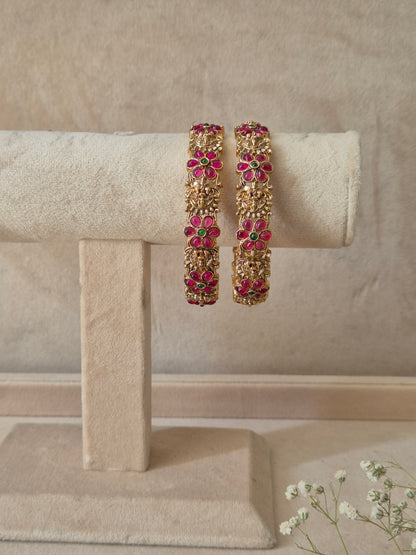  A set of two gold-plated Urmi bangles featuring intricate floral and textured metalwork inlaid with red and green gemstones, by naitri.in