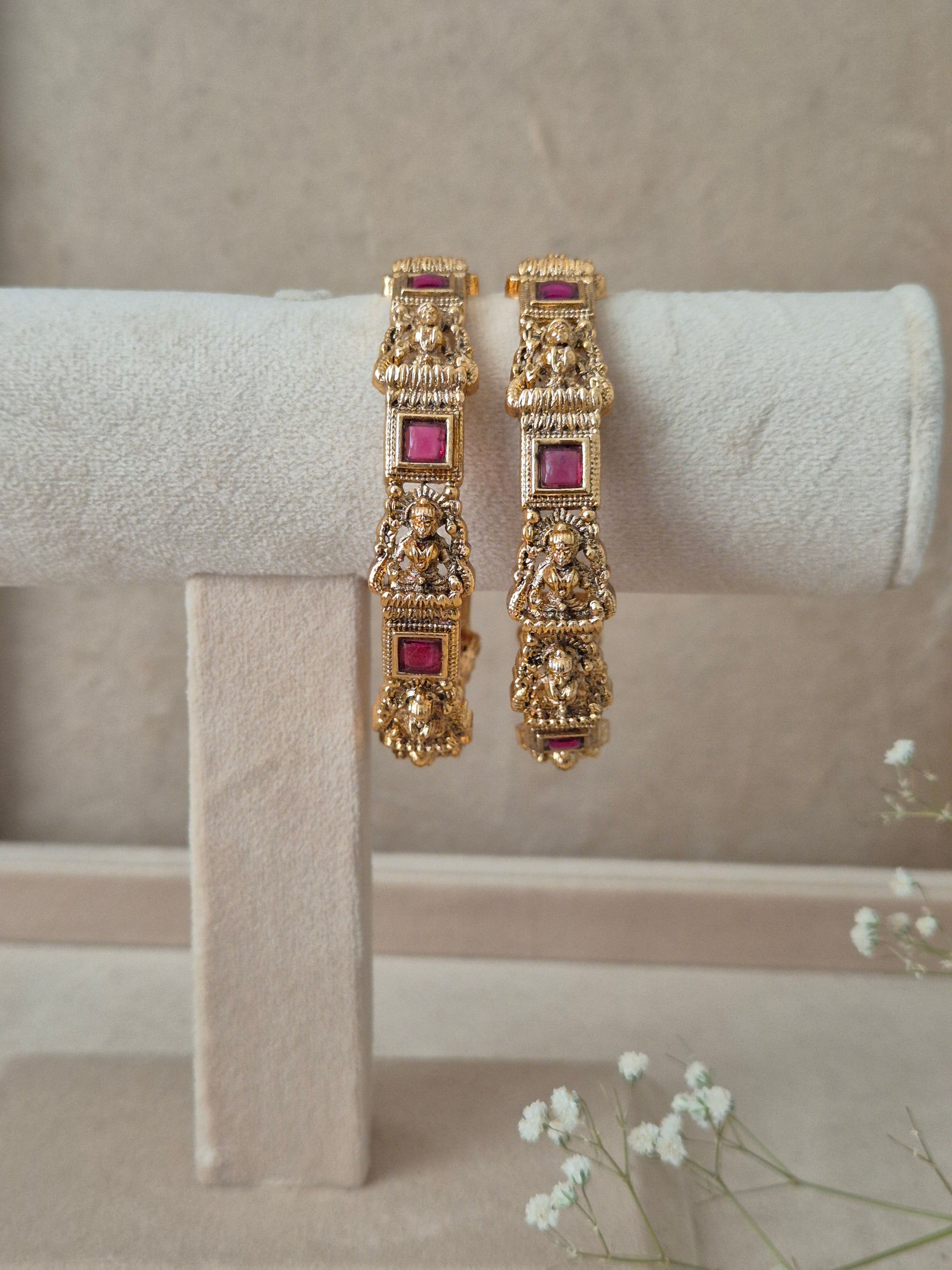 Set of 2 Urmila gold bangles by Naitri featuring intricate traditional temple-style carvings of deities, accented with square ruby-red gemstones, by naitri.in
