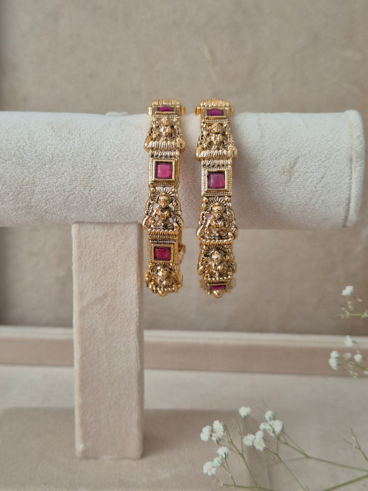 Set of 2 Urmila gold bangles by Naitri featuring intricate traditional temple-style carvings of deities, accented with square ruby-red gemstones, by naitri.in