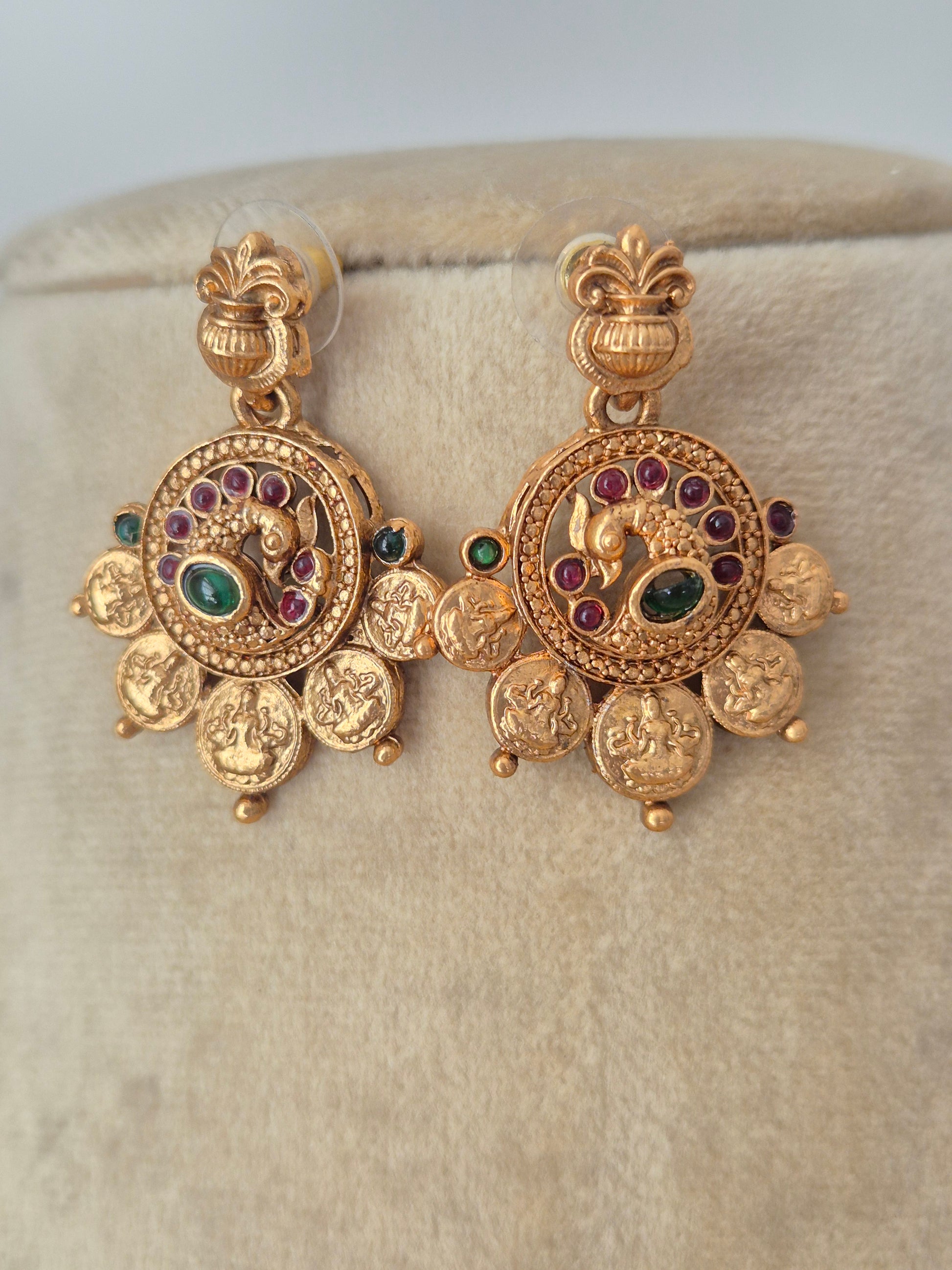 traditional Indian gold coin earrings with an antique finish, intricately carved floral and deity-style motifs, and small ruby-red and emerald-green stone embellishments by naitri.in