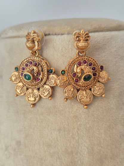 traditional Indian gold coin earrings with an antique finish, intricately carved floral and deity-style motifs, and small ruby-red and emerald-green stone embellishments by naitri.in