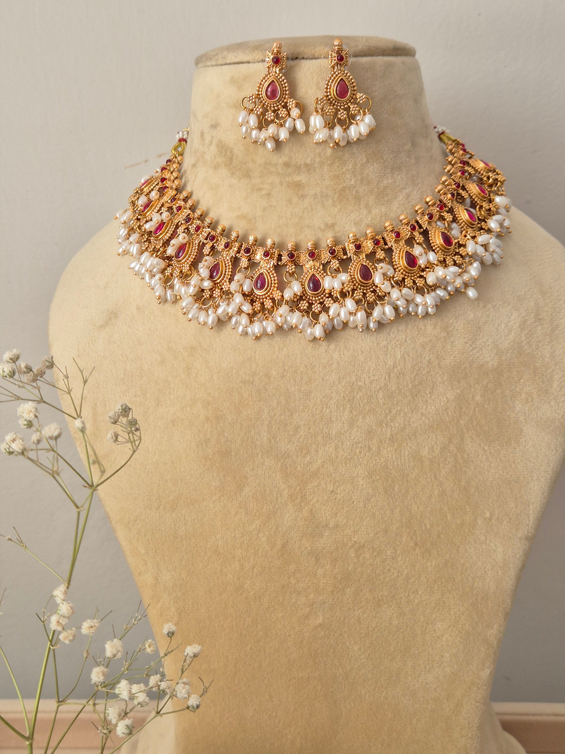 Handcrafted gold-plated chocker necklace featuring detailed traditional metalwork, ruby-red stone accents, and a rich white pearl cluster fringe, paired with matching teardrop drop earrings by naitri.in
