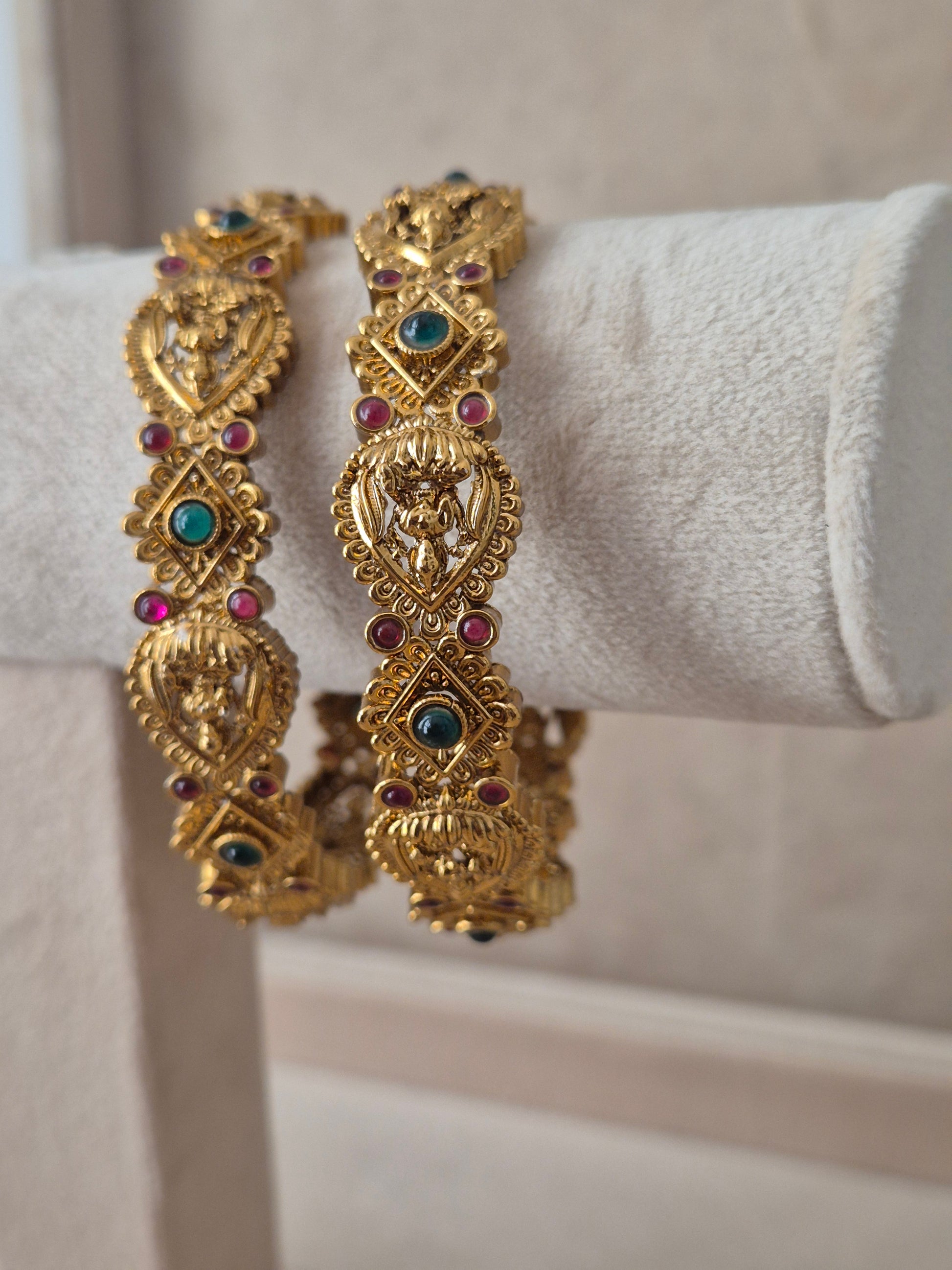 Pair of Naitri's Vaina gold bangles showcasing detailed ethnic craftsmanship, embedded with vibrant pinkish-red and green accent stones, by naitri.in