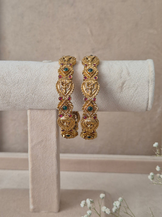 Set of 2 Vaina gold bangles by Naitri featuring intricate traditional filigree carvings, accented with alternating ruby-red and emerald-green gemstones by naitr.in