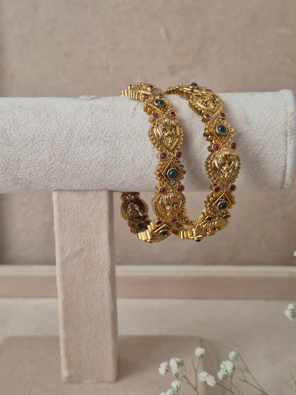 Two traditional gold-plated ethnic bangles with detailed textured patterns and inlaid red and green gemstones by naitri.in