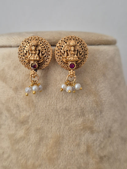 A pair of gold-toned Indian jhumka earrings with detailed Lakshmi motifs, small white pearl bead drops, and a central red gemstone, by naitri.in