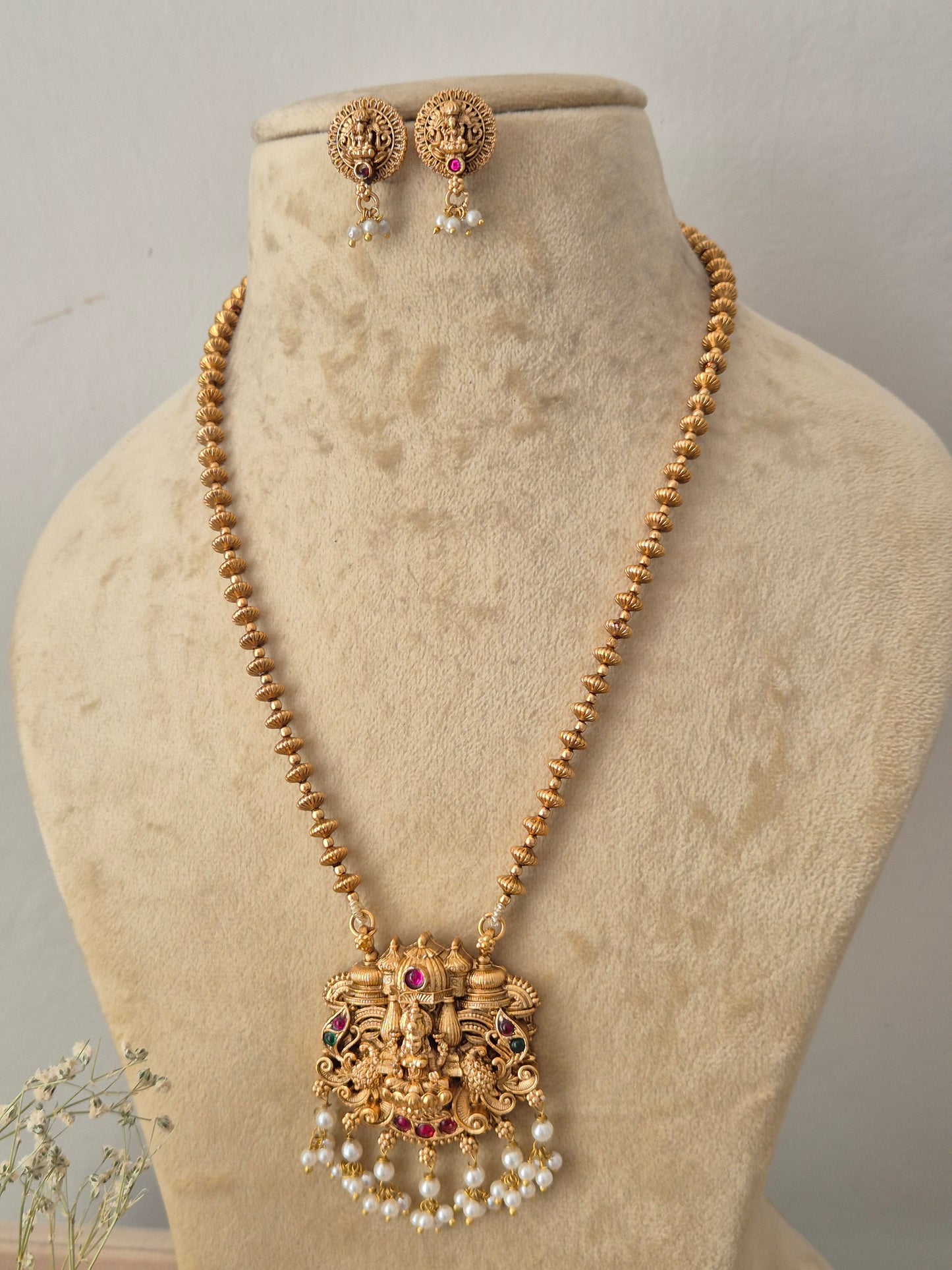 Gold-toned traditional Indian necklace and matching earrings. The necklace features a detailed filigree pendant with faux pearls and small red and dark accent stones for festive wear by naitri.in