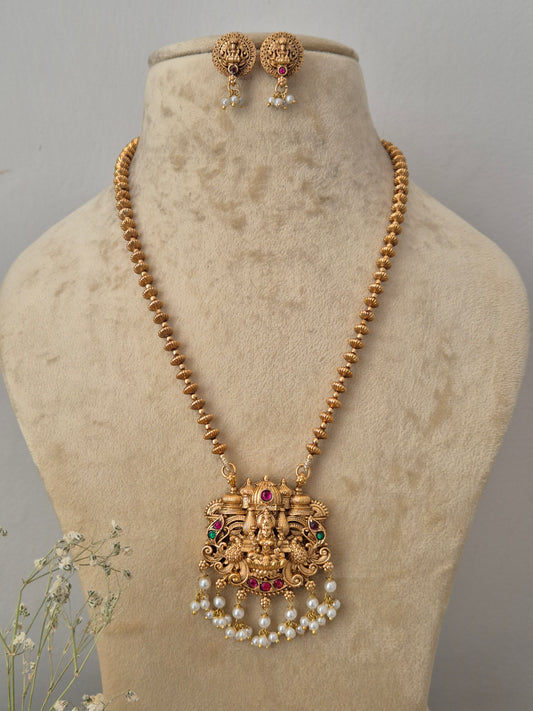 Gold-plated traditional Indian necklace set features a long, single strand of round gold-toned beads leading down to a large, ornate temple-style pendant engraved with a Lakshmi motif, accented with red and green gemstones and finished with a fringe of hanging white faux pearls. Matching round stud earrings with pearl droplets by naitri.in