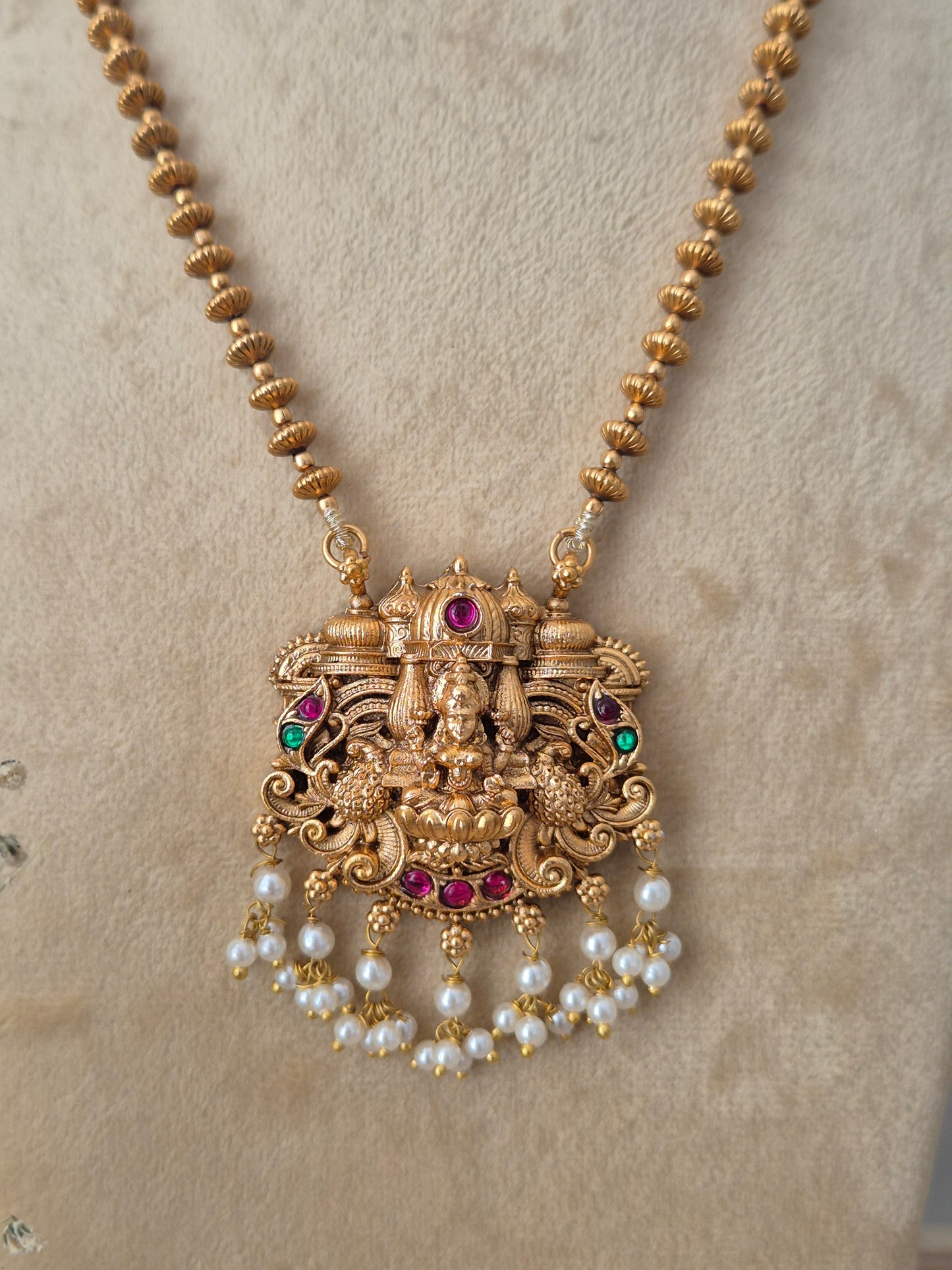 An ornate gold-toned traditional pendant necklace with a deity motif, accented with red and green stones and dangling pearl beads by naitri.in