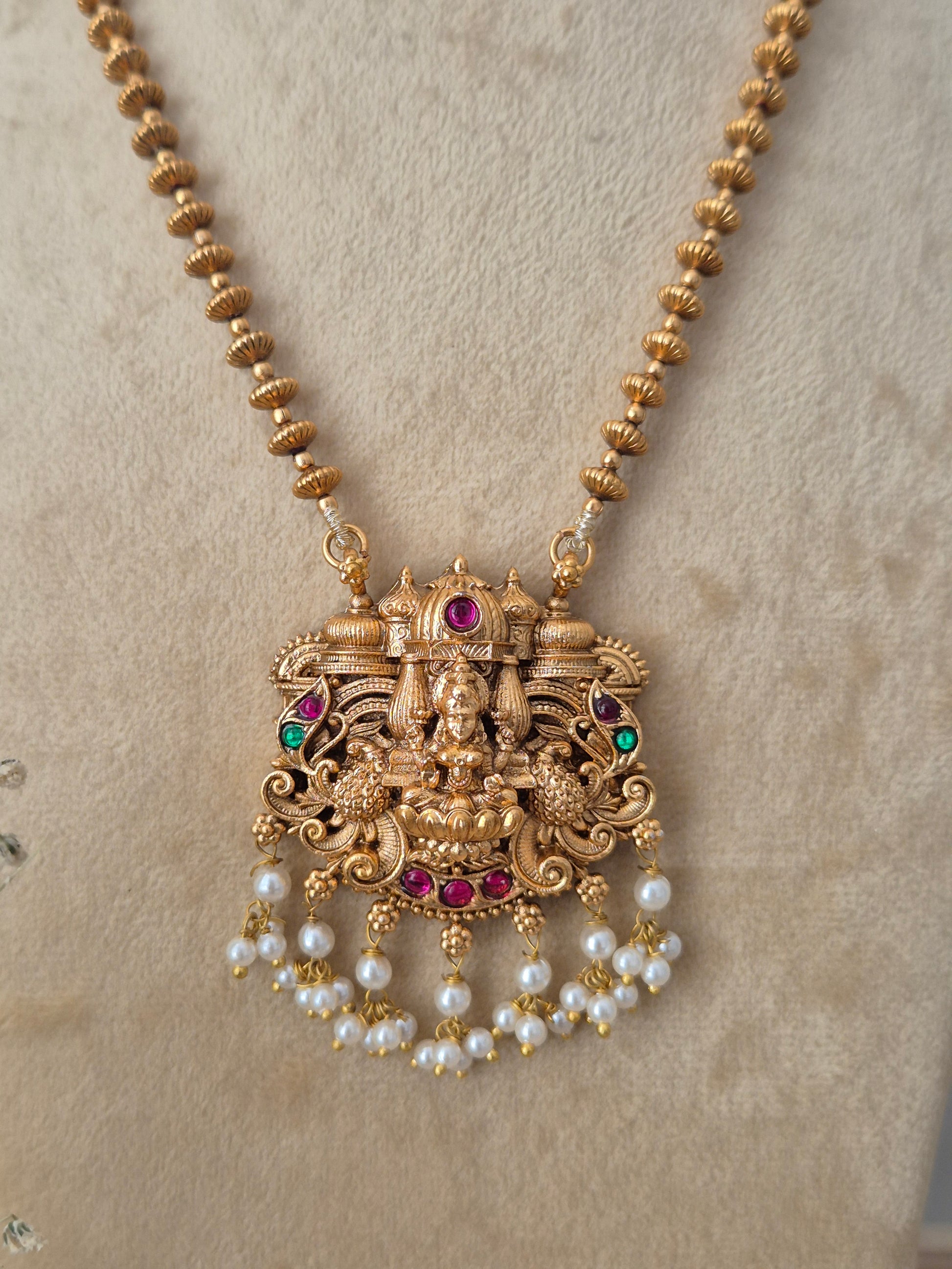 An ornate gold-toned traditional pendant necklace with a deity motif, accented with red and green stones and dangling pearl beads by naitri.in