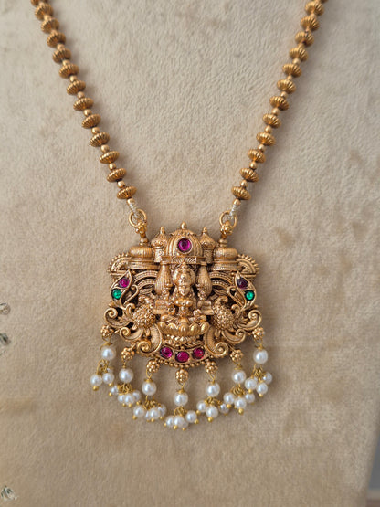 An ornate gold-toned traditional pendant necklace with a deity motif, accented with red and green stones and dangling pearl beads by naitri.in