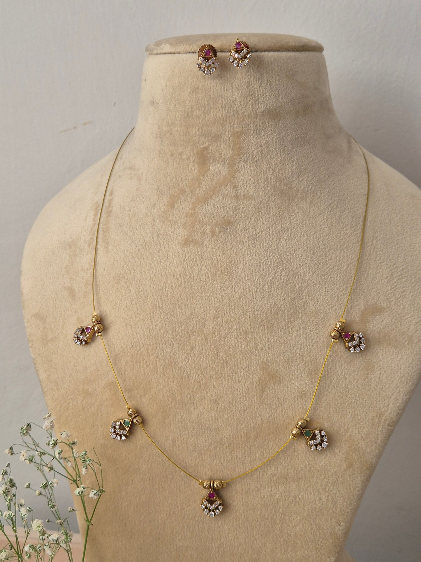 Delicate gold-toned fine chain necklace featuring five evenly spaced dangling ethnic charms accented with small pink and green gemstones, shown alongside matching stud earrings and dainty geometric floral drops by naitri.in