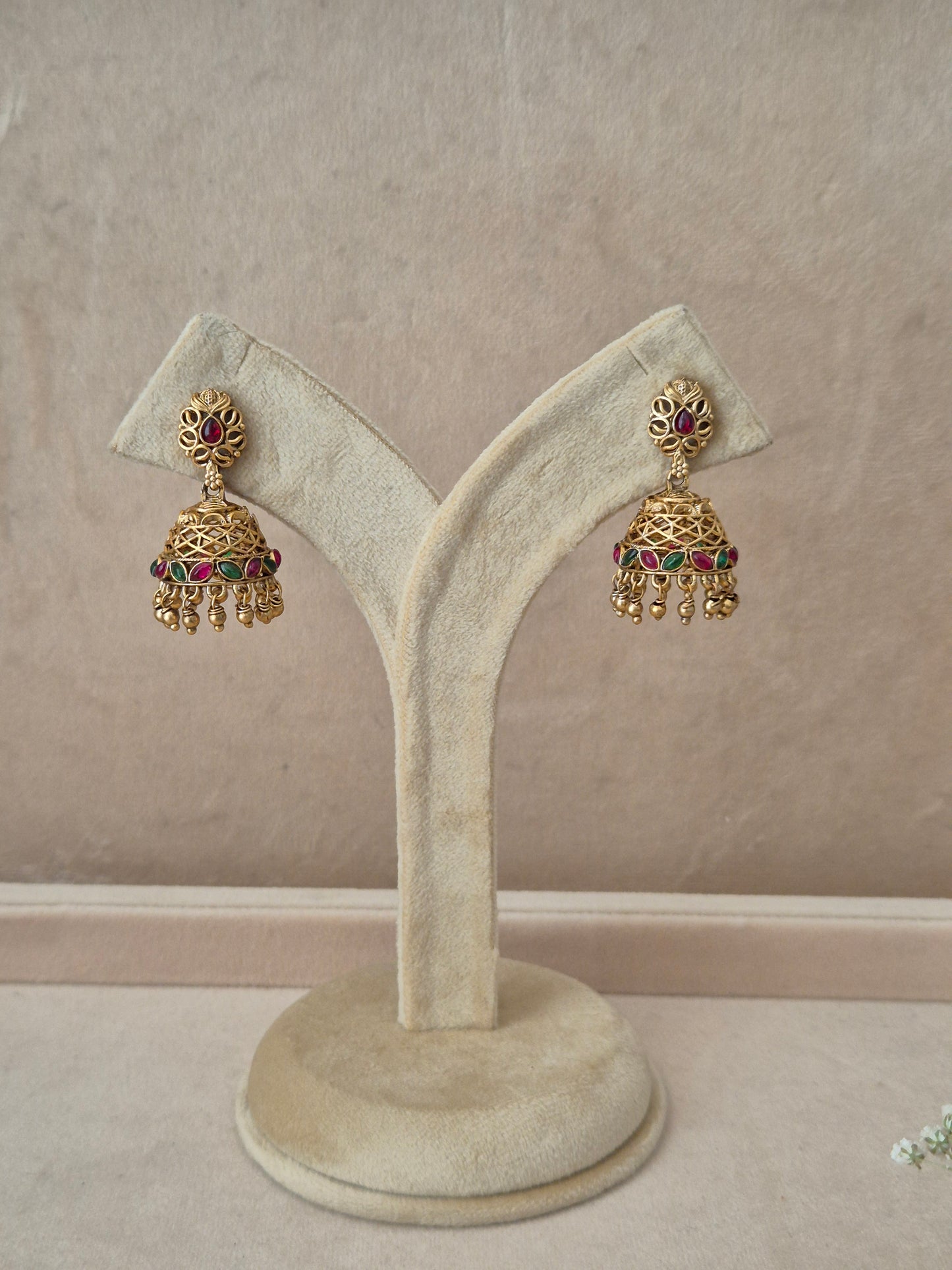 Pair of gold-plated Vedshri Jhumka earrings hanging on a beige velvet display stand, featuring floral-shaped studs with ruby red stones and detailed bell-shaped drops adorned with a band of green stones and small dangling gold beads by naiti.in