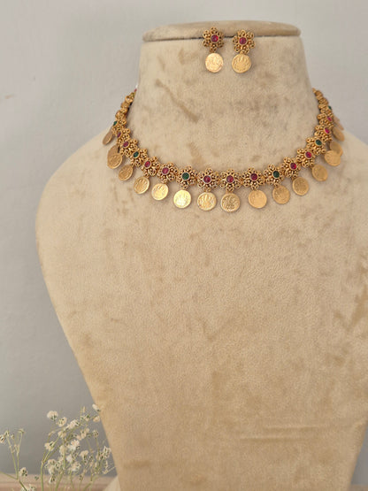 Gold-plated traditional Indian choker necklace and matching stud earrings features intricate floral motifs inset with tiny red and green gemstones, finished with a fringe of hanging circular gold coins, while matching coin stud earrings by naitri.in