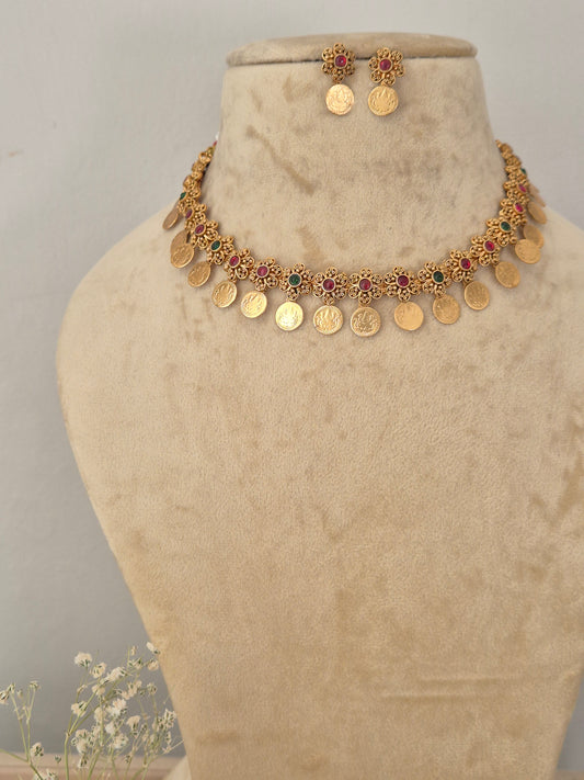 Gold-plated traditional Indian choker necklace and matching stud earrings features intricate floral motifs inset with tiny red and green gemstones, finished with a fringe of hanging circular gold coins, while matching coin stud earrings by naitri.in