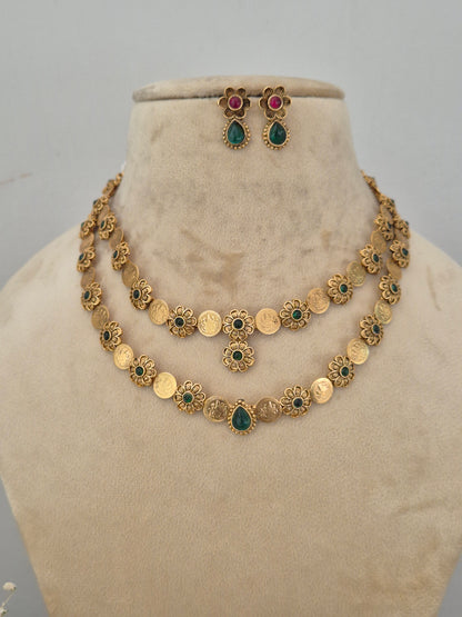 A gold-toned traditional necklace and matching earrings. The necklace features round gold coin-like charms, floral filigree details, and small green and red stone accents. by naitri.in