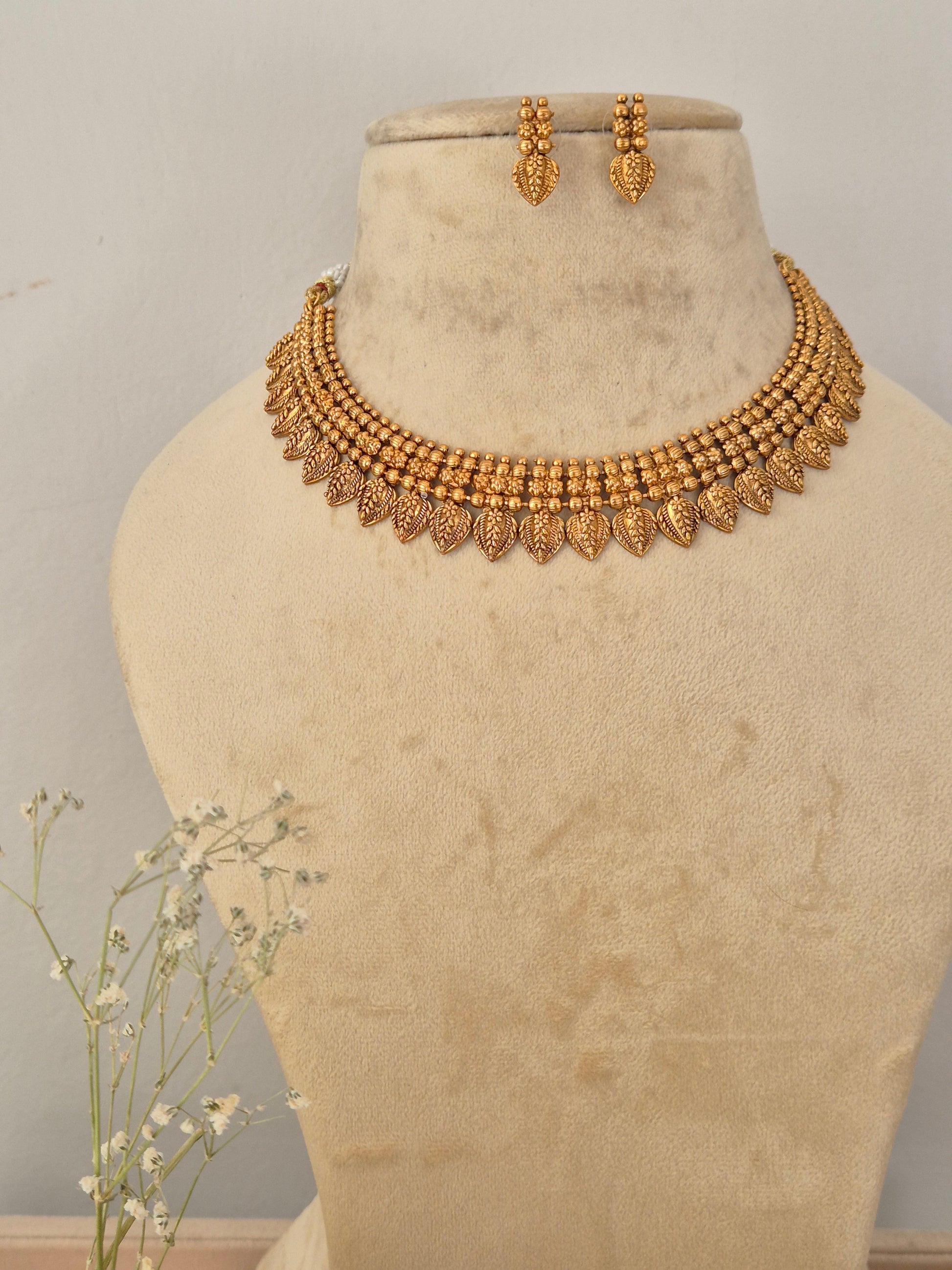 Gold-toned traditional Indian choker necklace and matching dangle earrings set the necklace features intricate, layered textured bead details and a lower fringe of detailed leaf-shaped metallic drops, by naitri.in