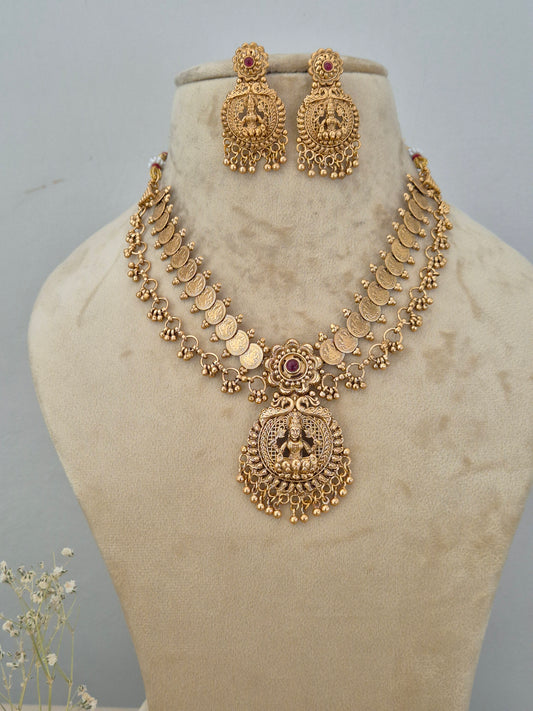 Gold-toned double-layered traditional temple jewelry set, the necklace features round coin-like charms with red gemstone accents and a large oval temple pendant featuring goddess motifs, paired with matching ornate drop earrings by naitri.in 