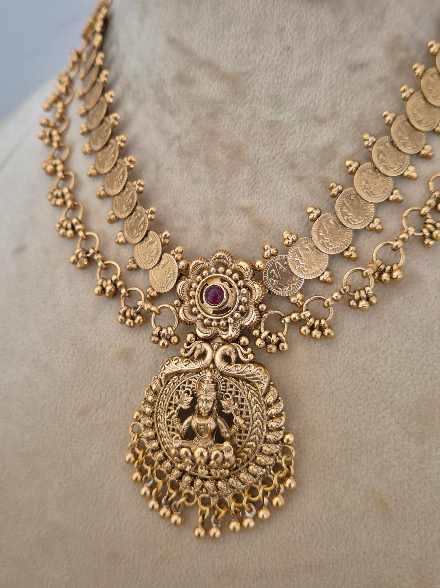 An ornate gold-toned traditional necklace with coin-shaped links, a central floral motif with a small purple stone, and a large detailed goddess pendant, by naitri.in
