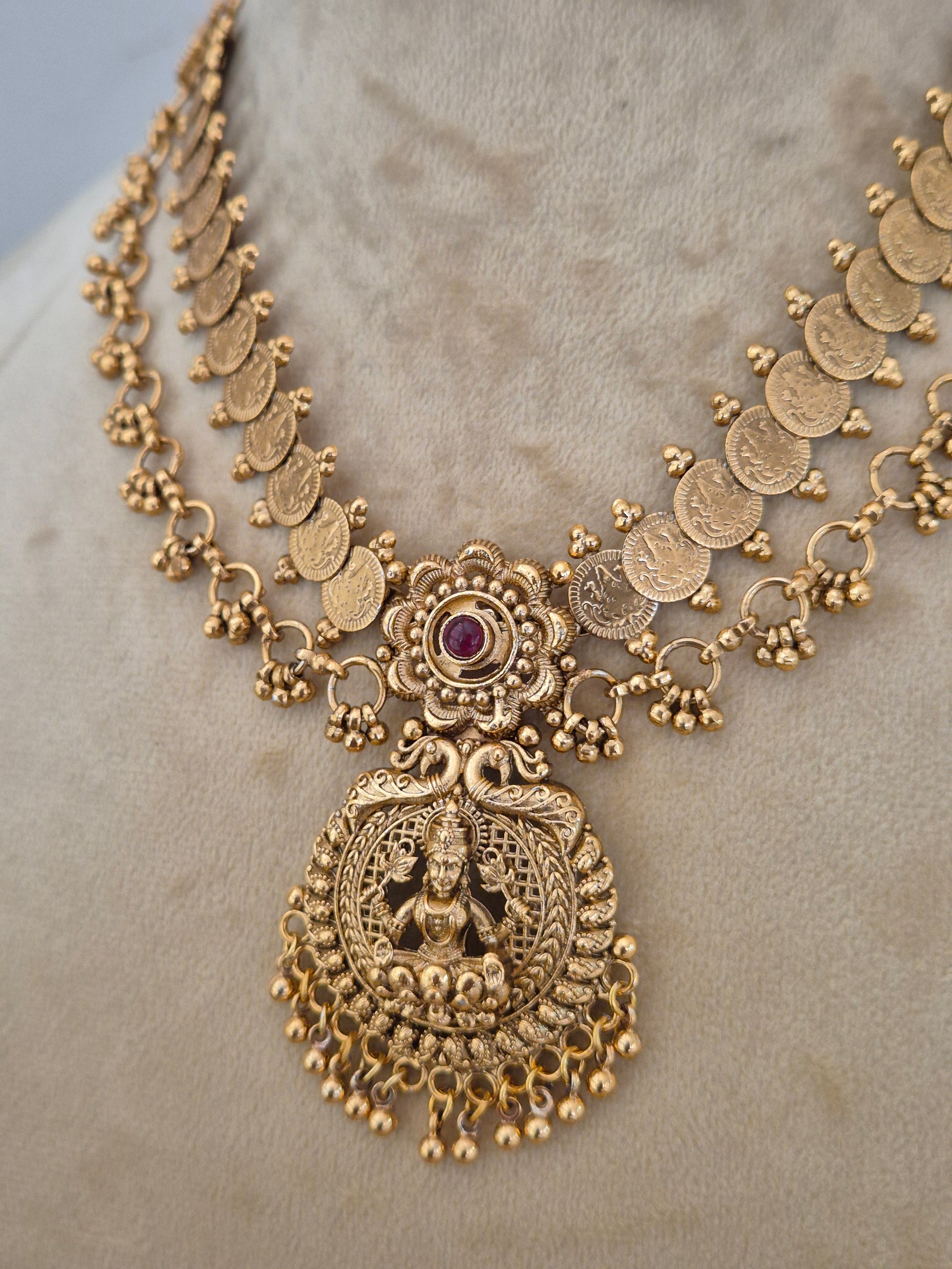 An ornate gold-toned traditional necklace with coin-shaped links, a central floral motif with a small purple stone, and a large detailed goddess pendant, by naitri.in