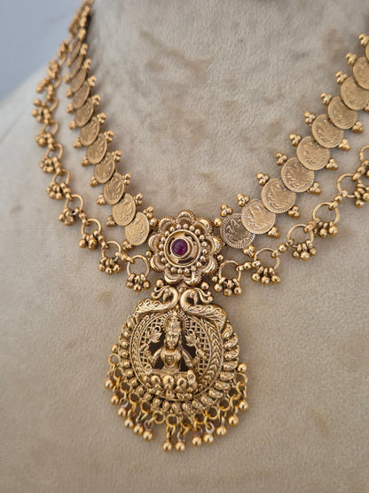 An ornate gold-toned traditional necklace with coin-shaped links, a central floral motif with a small purple stone, and a large detailed goddess pendant, by naitri.in