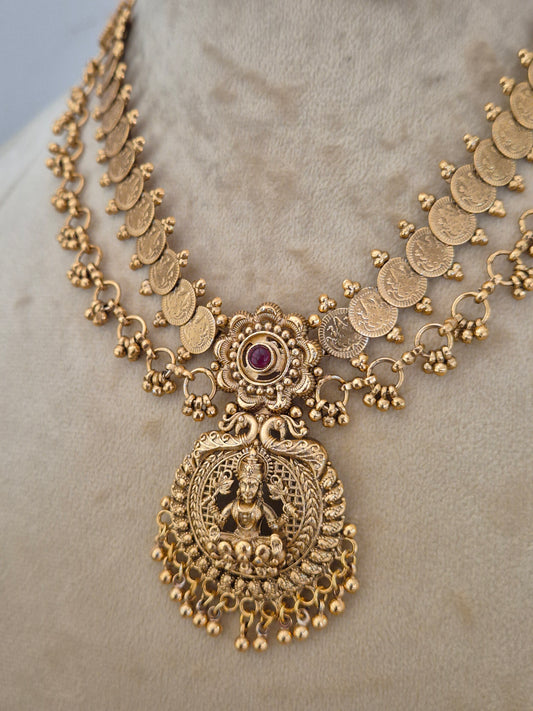 An ornate gold-toned traditional necklace with coin-shaped links, a central floral motif with a small purple stone, and a large detailed goddess pendant, by naitri.in