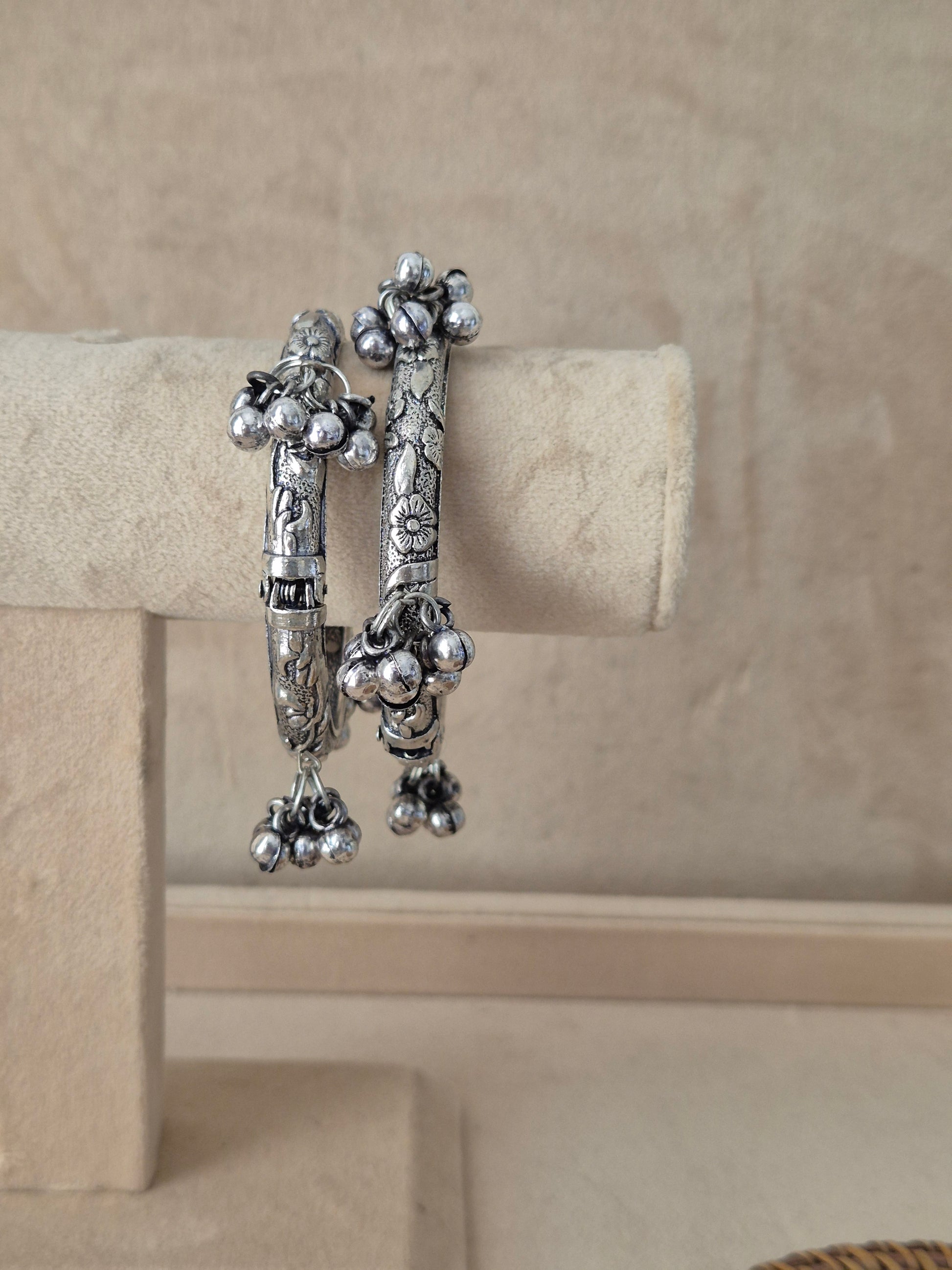 Set of two oxidized silver Wishi bangles hanging on a beige fabric display stand, featuring detailed embossed floral vine patterns and a cluster of dangling tribal jhumka charms with tiny metallic beads by naitri.in