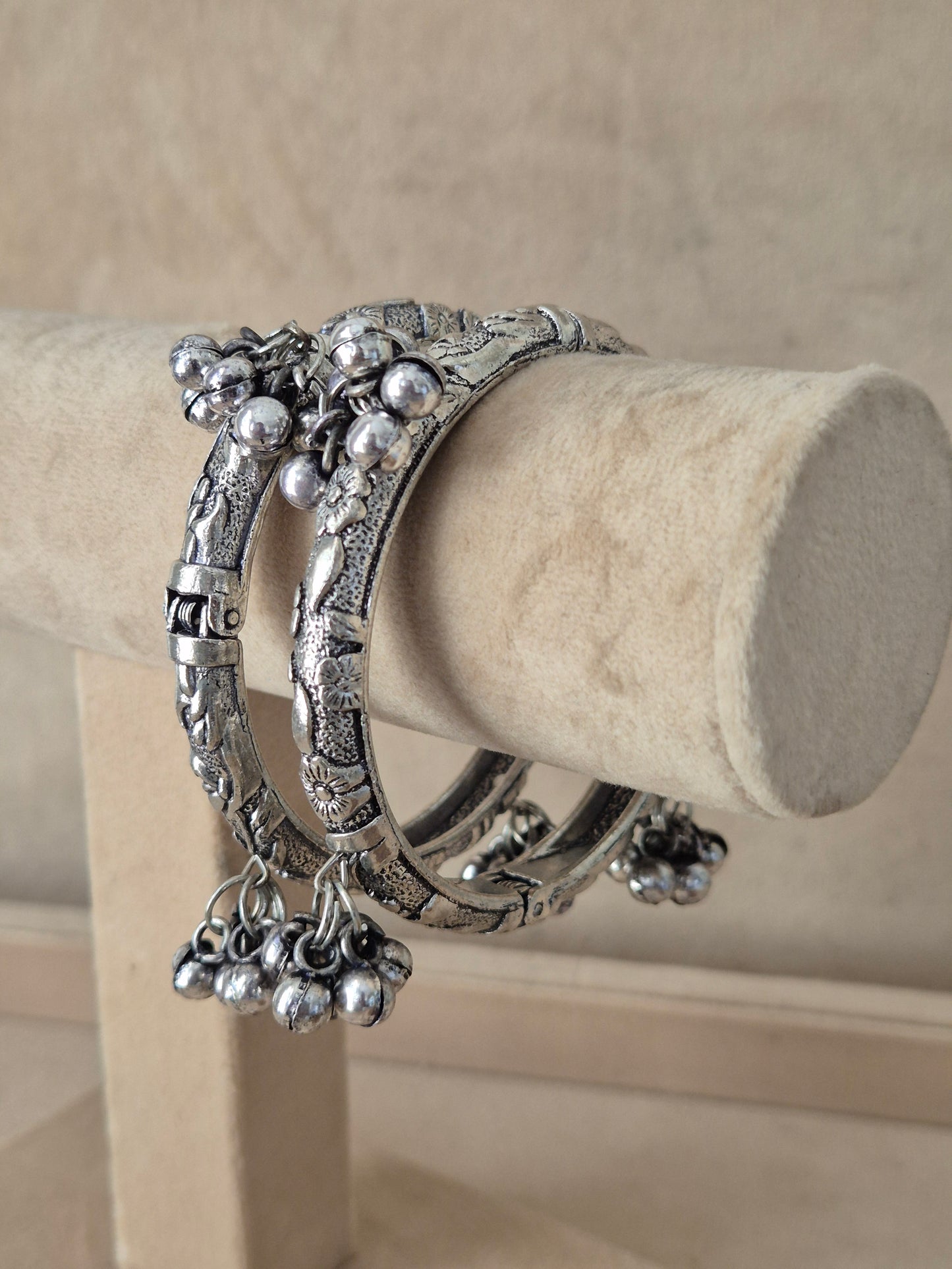 Statement mixed-metal bohemian Kada bangle set featuring intricate floral cutout textures and hanging ghungroo accents, exhibited on a luxury jewelry display holder by naitri.in