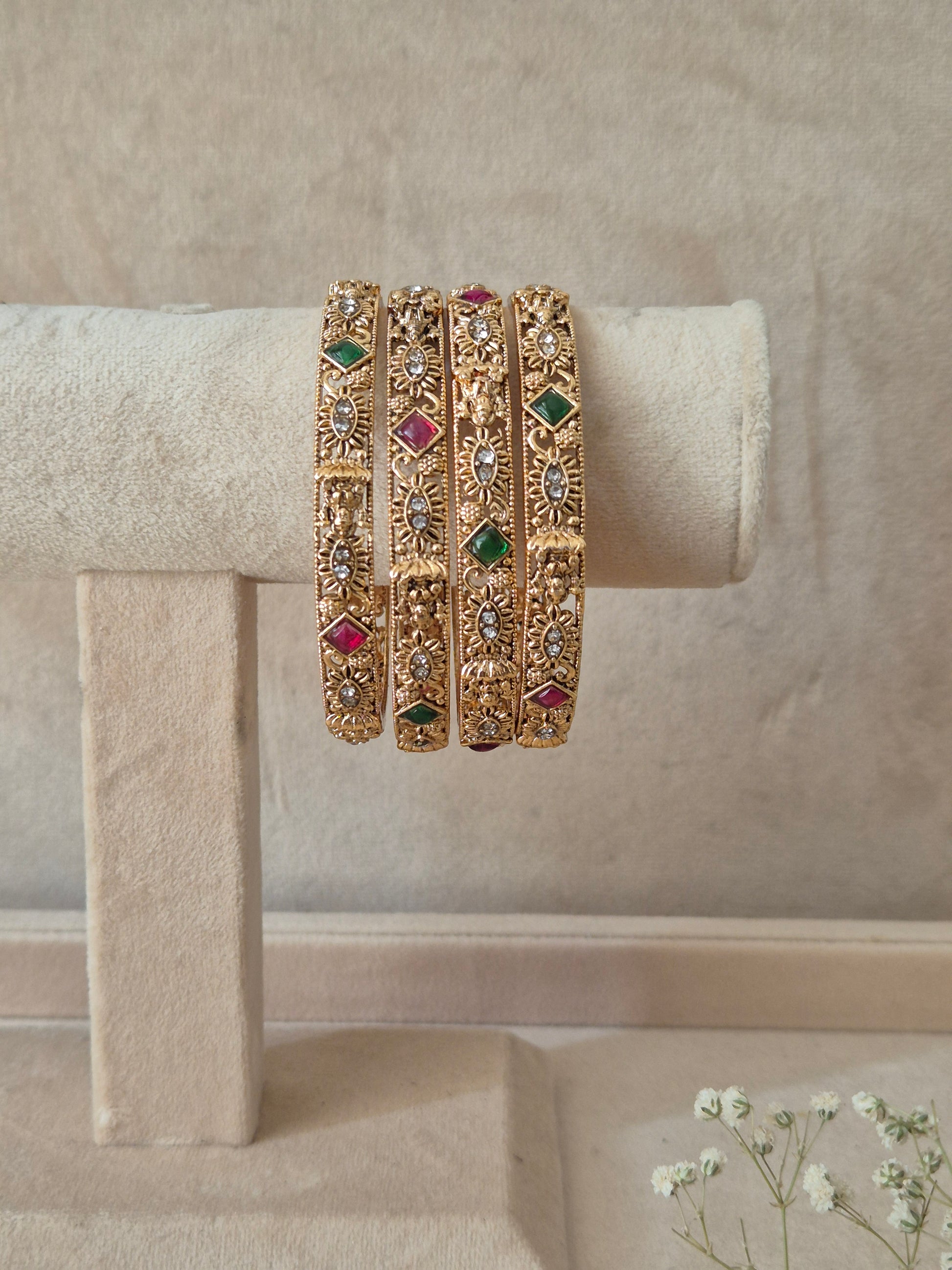 A set of four gold-plated Yamini bangles featuring intricate floral and textured metalwork inlaid with alternating red and green gemstones by naitri.in