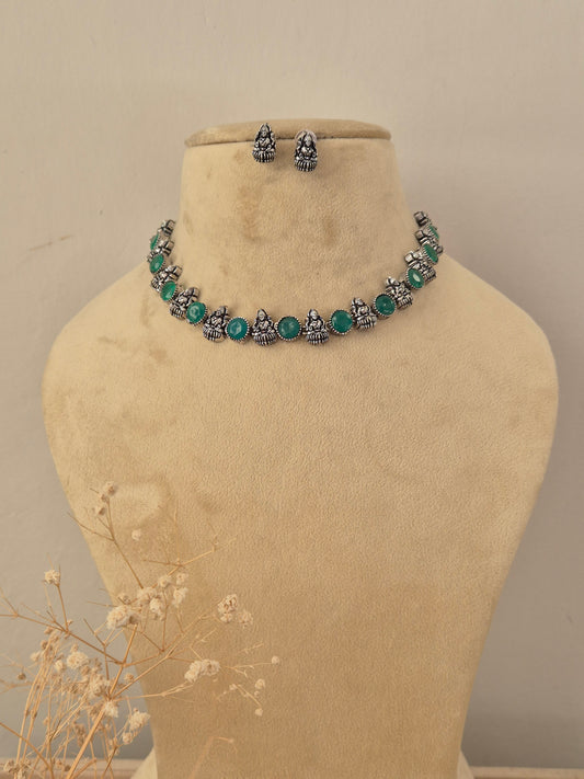 A silver-toned jewelry set  featuring a choker necklace and matching earrings with small Buddha figurines and round green gemstone accents by naitri.in
