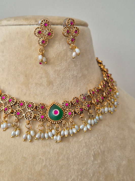 Intricately crafted antique gold choker set with vibrant ruby pink floral clusters, an emerald green center stone, and delicate pearl fringe detailing by naitri.in