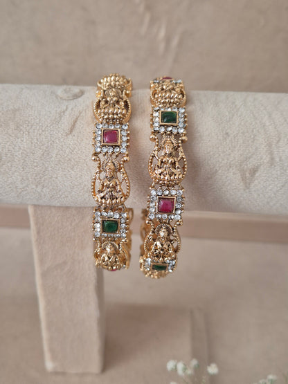 Two traditional gold-plated ethnic bangles with detailed religious carvings and inlaid red and green gemstones By naitri.in