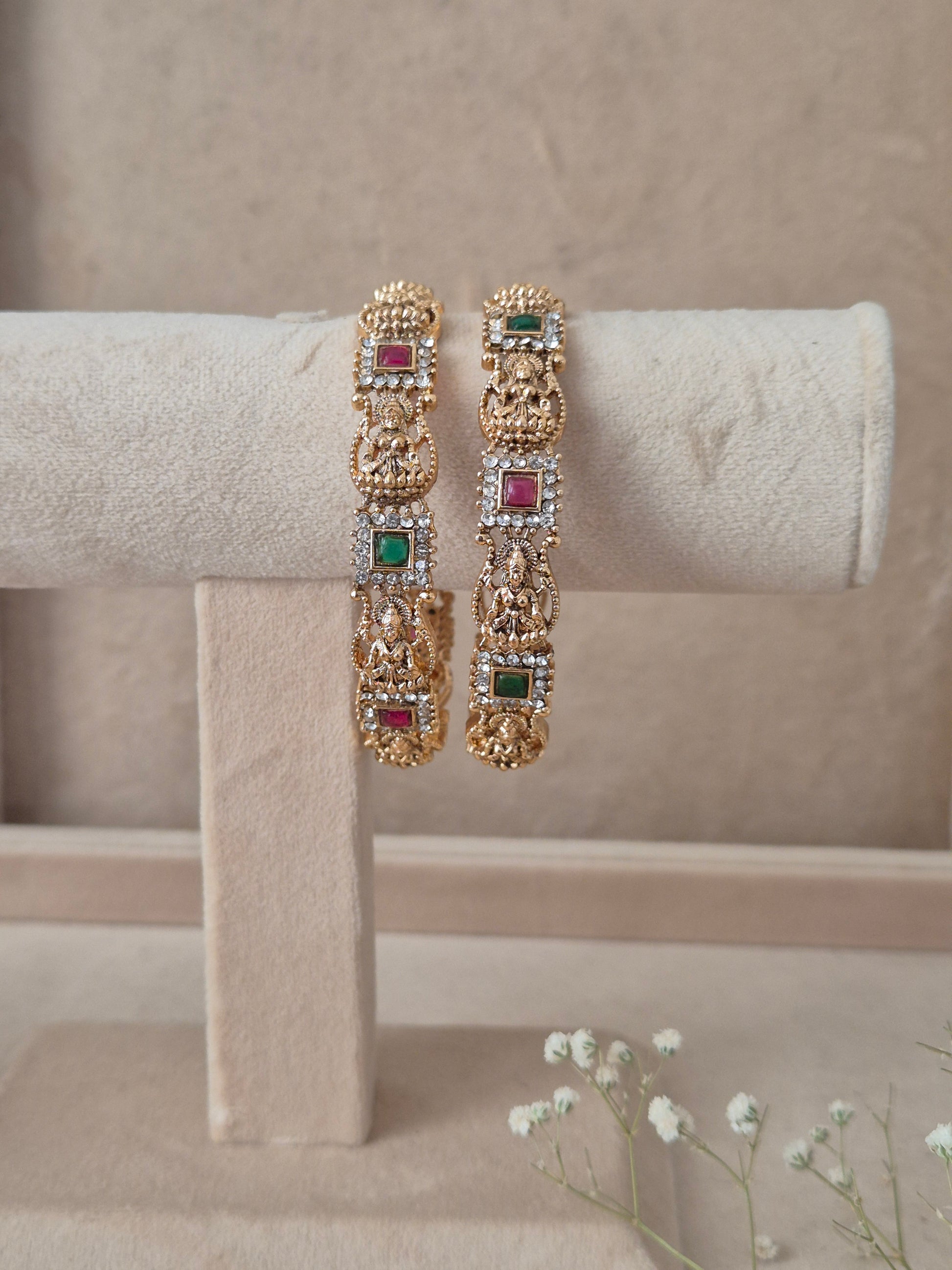 Set of 2 Yogshi gold bangles by Naitri featuring intricate traditional temple-style deity carvings, accented with alternating ruby-red and emerald-green gemstones, by naitri.in