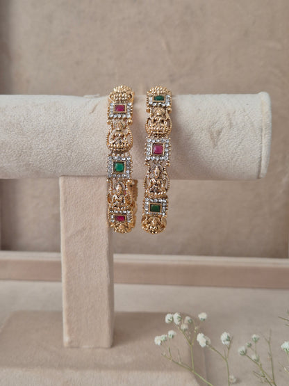 Set of 2 Yogshi gold bangles by Naitri featuring intricate traditional temple-style deity carvings, accented with alternating ruby-red and emerald-green gemstones, by naitri.in
