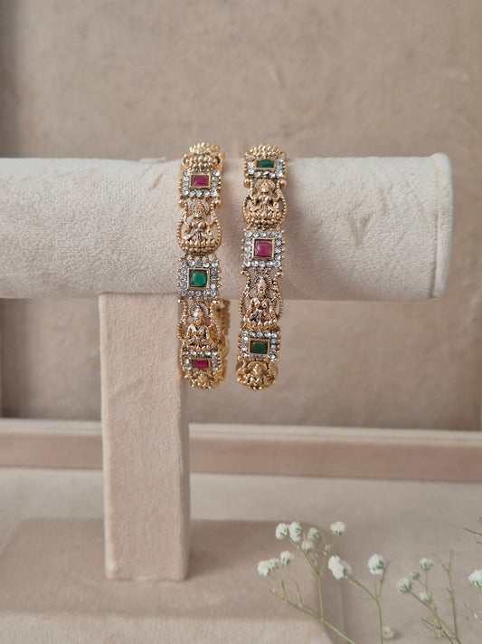 Set of 2 Yogshi gold bangles by Naitri featuring intricate traditional temple-style deity carvings, accented with alternating ruby-red and emerald-green gemstones, by naitri.in
