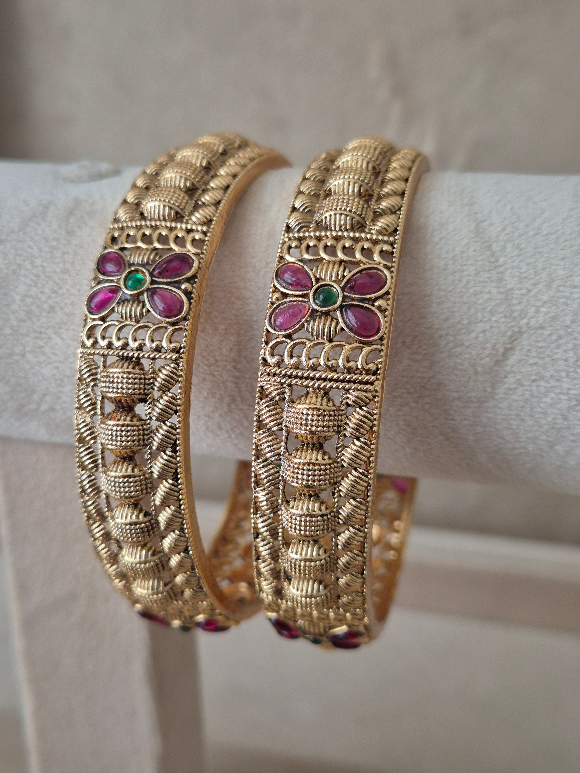 Two traditional gold-plated ethnic bangles with detailed geometric textures and inlaid red and green gemstones by naitri.in