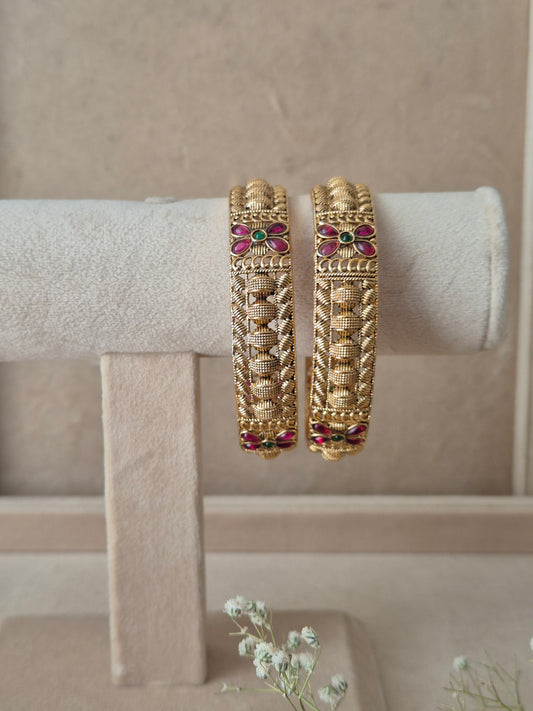 Set of 2 Zahira gold bangles by Naitri featuring intricate traditional filigree and leaf patterns, accented with ruby-red and emerald-green floral gemstones by naitri.in