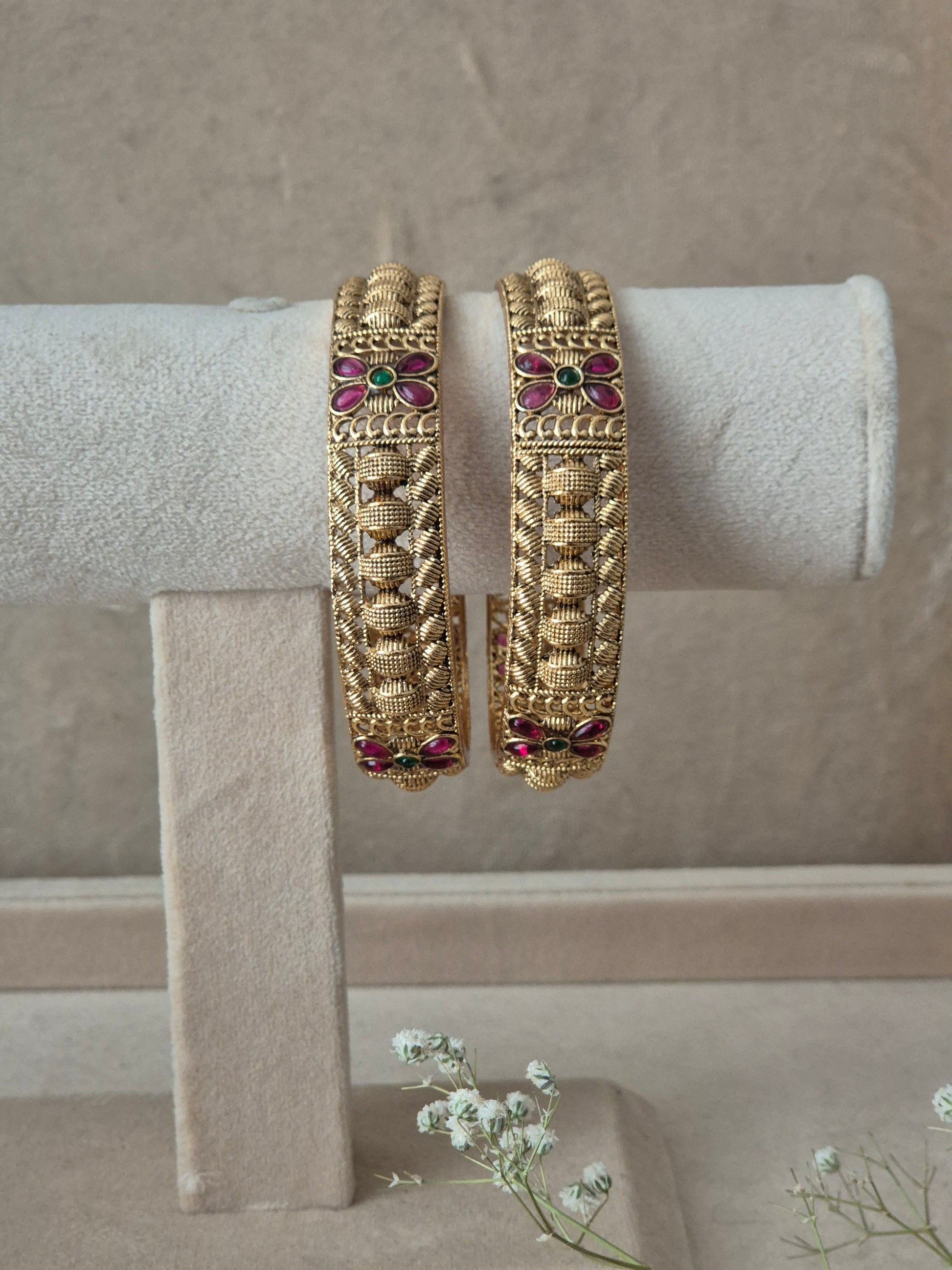 Pair of Naitri's Zahira gold bangles showcasing detailed antique-style craftsmanship, embedded with vibrant pinkish-red floral motifs and green accent stones, by naitri.in